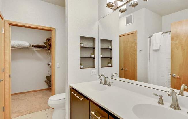 A modern bathroom here at Blue and Lime featuring a wide double vanity with a smooth countertop, dual sinks, and sleek brushed metal fixtures beneath a large wall mirror with overhead lighting. Built-in recessed shelves add decorative storage, while light tile flooring, a shower with a white curtain, and a walk-in closet create a clean, functional layout.
