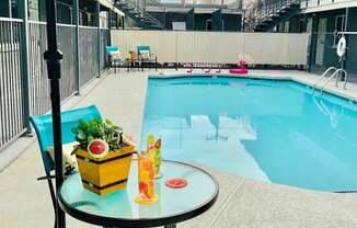 A table with a yellow container and a glass of orange juice is on a patio next to a pool.