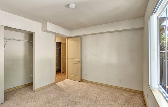 A comfortable bedroom here at Asbury Park featuring soft carpet flooring, neutral-toned walls, and a large window that brings in natural light, complemented by an open closet with shelving, a nearby hallway with wood-style flooring, and ample wall space suitable for a bed, dresser, or additional storage.