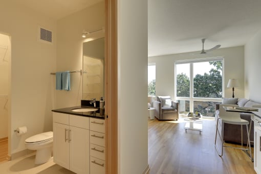 Glimpse into Flat B studio apartment floor plan at Coze Flats, bathroom has black granite counter, white cabinets. Past the bathroom is the living and sleeping area with oversized, East facing windows with treetop view