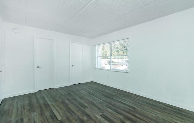 A spacious, empty room with light-colored walls and a textured ceiling. It features two closed doors on the left side and a large window on the right, allowing natural light to illuminate the space. The floor is covered with brown laminate flooring, creating a modern and clean aesthetic.
