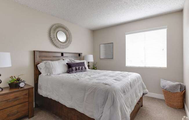 A bedroom with a large bed, a nightstand, a clock, a plant, a mirror, and a window.