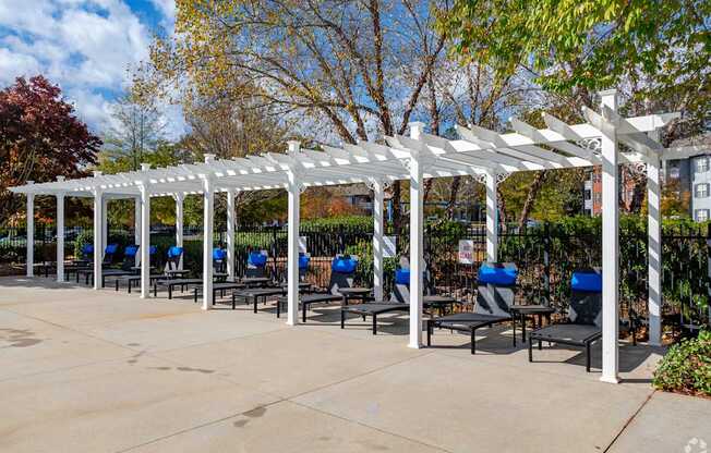 A white pergola with blue chairs is in the foreground of a sunny day.