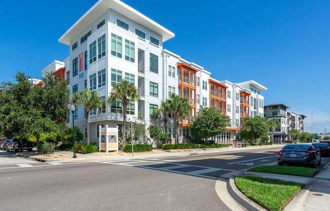 A modern multi-story building with a white and orange facade is situated on a street corner with cars parked along the curb.