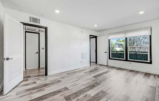 A room with white walls and wooden flooring with a view of trees outside the window.
