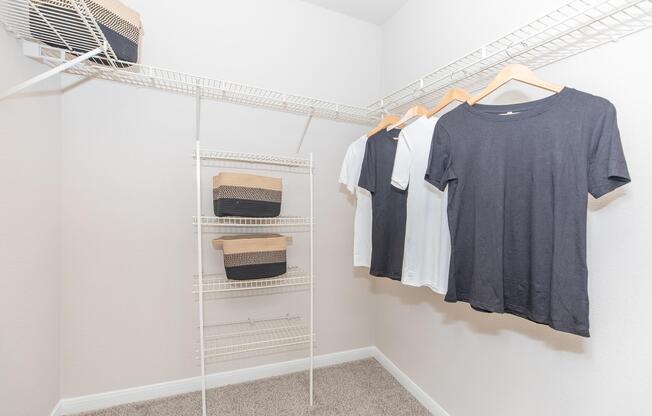 A clean, organized closet featuring a white wire shelving unit. Hanging on the left are several shirts in black and white, while below are stacked woven baskets in a contrasting color. The walls are painted in a light shade, and the floor is carpeted.