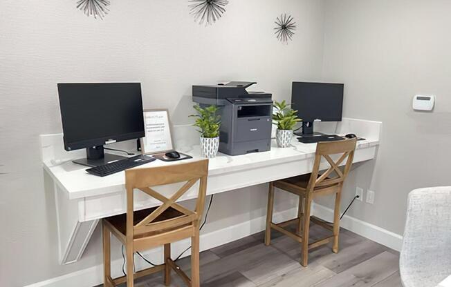 A modern shared workspace featuring two computers with monitors and keyboards placed on a sleek white desk. A printer is located on the desk, alongside two potted plants. Two wooden chairs are positioned at the desk. The wall is decorated with minimalist wall art and there's a sign on the desk.