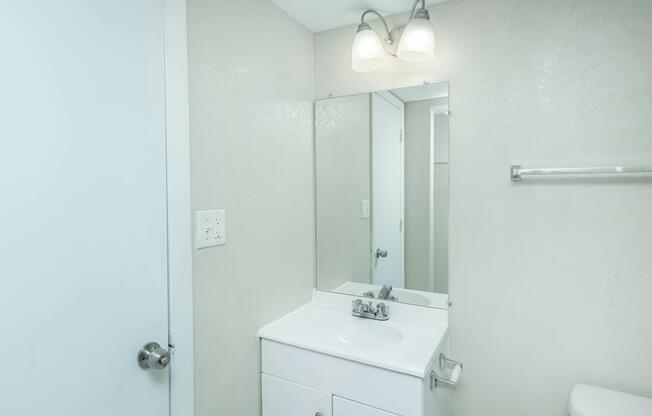 A clean and simple bathroom featuring a white vanity with a sink and faucet, a large mirror above the sink, a light fixture with two bulbs, and a white toilet. The walls are painted a light gray, and there is a towel bar mounted on the wall. A door leads out of the bathroom.