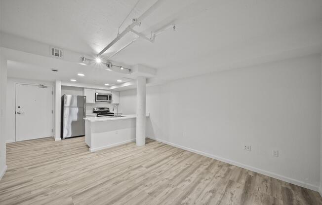 A bright, open-concept living space featuring light-colored walls and wood laminate flooring. The kitchen includes stainless steel appliances, a countertop, and bar seating. Natural light flows into the area, which is devoid of furnishings, creating a spacious, inviting atmosphere.