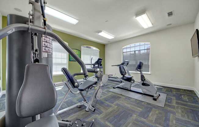 A bright community fitness center here at Mission Green featuring a well-equipped workout space with cardio machines, strength training equipment, patterned flooring, large windows with blinds for natural light, soft green accent walls, and overhead lighting that creates an open and energizing environment.