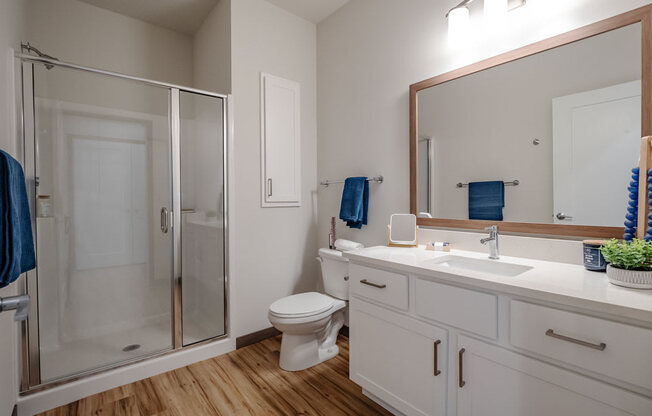 A modern bathroom here at Van Buren East with a glass-enclosed shower and white vanity. A large mirror, bright lighting, and wood-style flooring create a clean, fresh, and inviting atmosphere.