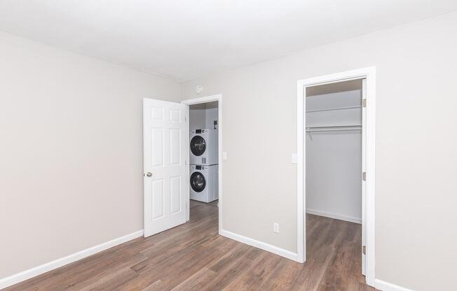 A small, light-colored room featuring a door on the left leading outside, a door on the right revealing a closet, and a wall-mounted washer and dryer in the back. The flooring is a warm wooden color, and the walls are painted in a soft, neutral tone, creating a bright and spacious feel.