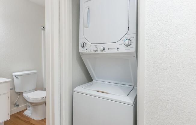 A compact laundry area featuring a stacked washer and dryer unit beside a toilet. The space has light-colored walls and wooden flooring, creating a clean and organized appearance.