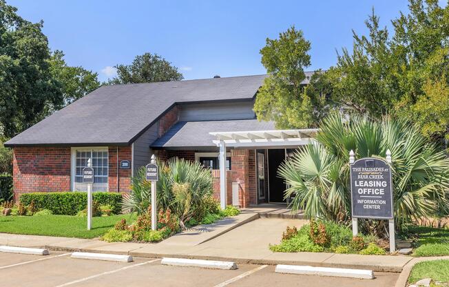 A brick building with a gray roof, featuring a leasing office sign and landscaping with palm trees and green bushes. The entrance has a covered walkway, and the parking lot is visible. The sky is clear and blue, indicating a sunny day.