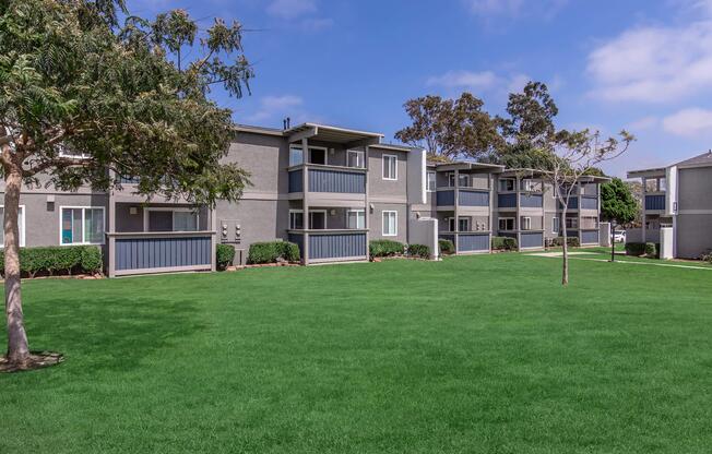 A view of a residential apartment complex featuring two-story buildings with balconies, set against a bright blue sky. Lush green grass and a few trees are visible in the foreground, creating a welcoming outdoor space.