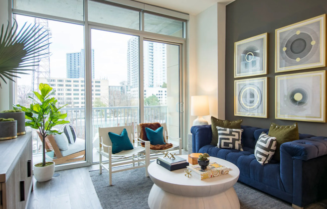 An spacious living room with a couch, two armchairs, a coffee table, and a sideboard. The room has dark wood-style floors, full-width floor-to-ceiling windows, a dark gray accent wall, a balcony, and a view of the city skyline.