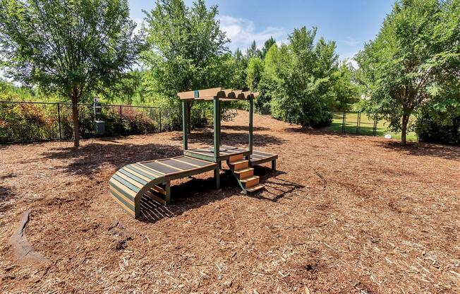 Gated dog park with mulch, trees, and a piece of agility equipment with steps and a raised surface