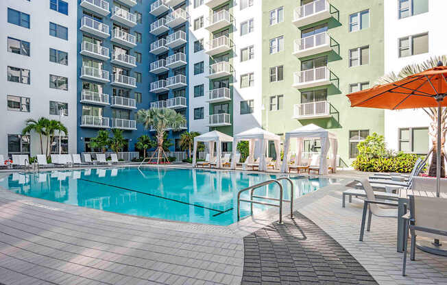 A large swimming pool surrounded by lounge chairs and umbrellas in front of apartment buildings.