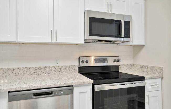 A modern kitchen with white cabinets and a granite countertop. It features a stainless steel microwave above an electric stove and oven, along with a sleek dishwasher. The backsplash is a white tile, creating a clean and vibrant look. Ideal for cooking and entertaining.