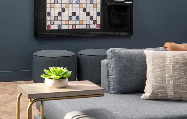 A living room with a grey couch, a small table with a plant and a book, and a framed board game on the wall.