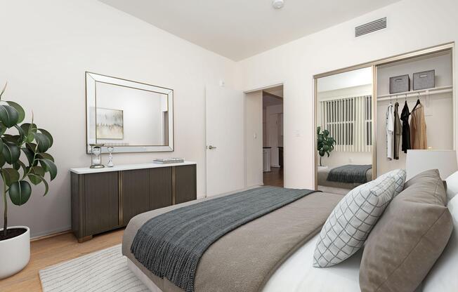 A modern bedroom featuring a neatly made bed with gray and white bedding, a decorative throw blanket, and a textured pillow. There is a potted plant in the corner, a mirrored wardrobe reflecting the room, and a dresser with a large mirror above it. The walls are light-colored, creating a spacious and inviting atmosphere.
