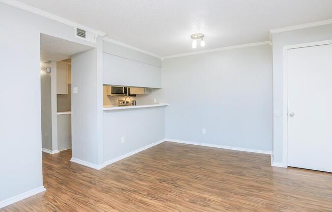 Light-filled interior of a modern apartment featuring grey walls and laminate flooring. The layout includes an open living area transitioning into a kitchen with a bar counter. A closed door leads to another room, and the space is illuminated by a ceiling light fixture.