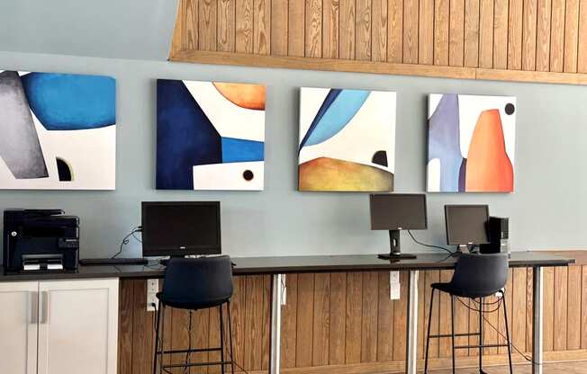 a row of stools sit in front of a desk with a computer and three paintings on