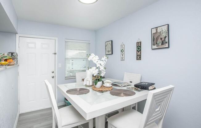 A bright dining area featuring a white table with four matching chairs, set with placemats and a magazine. A window with blinds allows natural light, while a door leads outside. The walls are painted light blue, adorned with framed artwork and a vase of white orchids on the table, creating a cozy and inviting atmosphere.
