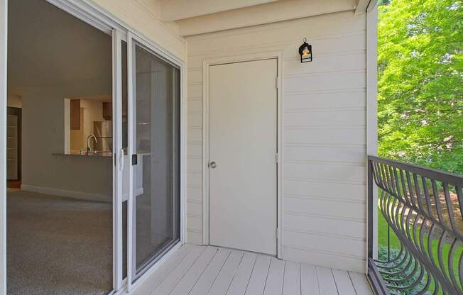 A private outdoor balcony here at Adagio featuring light painted decking, a decorative metal railing, and a sliding glass door that connects to the living area, with an exterior storage closet and wall-mounted light fixture, while leafy trees and landscaped greenery beyond the railing create a peaceful outdoor extension for relaxing or enjoying fresh air.