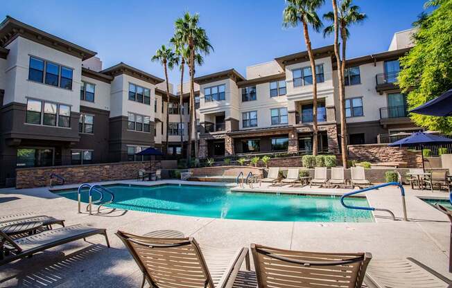 A swimming pool surrounded by lounge chairs and palm trees in front of a multi-story apartment building.