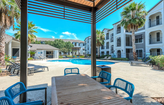 A wooden table is in the foreground of a sunny poolside area.