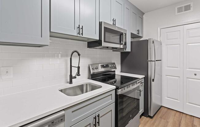 Modern kitchen featuring light blue cabinets, stainless steel appliances, including an oven, microwave, and refrigerator. A single basin sink with a stylish black faucet is situated on a white countertop. The kitchen has white subway tile backsplash and a door leading to another room.