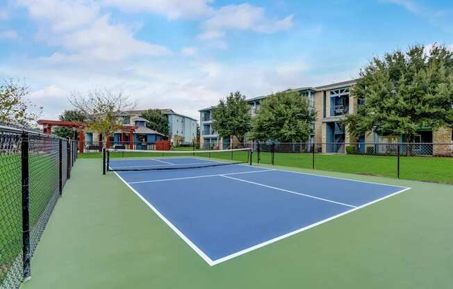 A tennis court is surrounded by a black fence and a building is in the background.