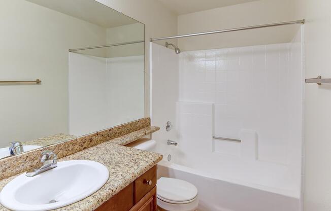 A clean bathroom featuring a bathtub with a shower, a white sink with a granite countertop, and a large mirror above the sink. The walls are light-colored, and there is a towel bar on the side. The overall design is modern and minimalistic.
