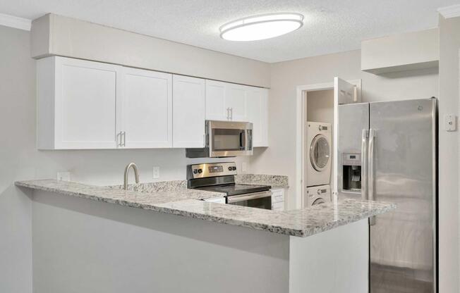 Modern kitchen featuring white cabinets, stainless steel appliances, a granite countertop, and an overhead light fixture. The layout includes a stove, microwave, and refrigerator, with an adjoining space for a washer and dryer visible in the background.