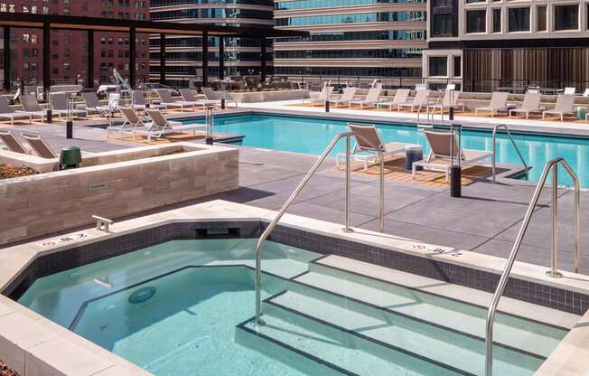 A pool with a hot tub and chairs in front of a cityscape.