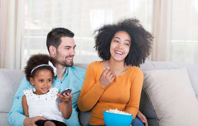 A family of three is sitting on a couch, with the woman holding a bowl of popcorn and the girl holding a remote control.