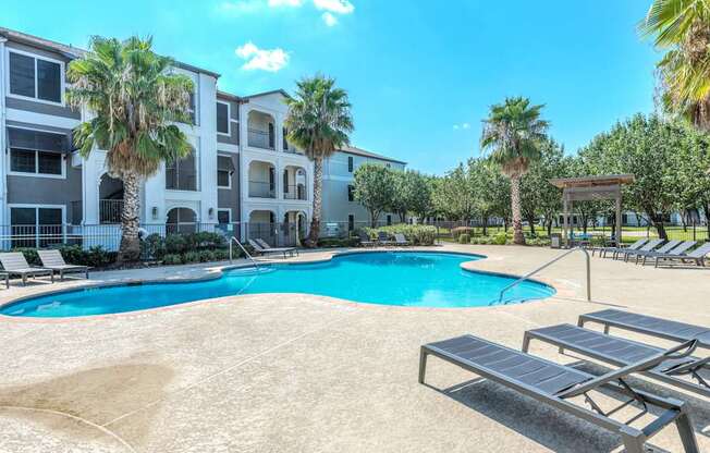 A swimming pool surrounded by sun loungers and palm trees in front of a multi-story apartment building.