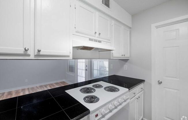 Modern kitchen featuring white cabinetry, a black countertop, and a stovetop with four burners. There's a range hood above the stove, and the wall behind it is partially visible. A door on the right leads to another room, and large windows create a bright atmosphere. The flooring is light-colored wood.