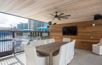 An open-air dining room by the pool enclosed with a gate, with a wood-paneled wall and ceiling, a flat-screen TV mounted on the wall, ten white upholstered chairs, and a ceiling fan.