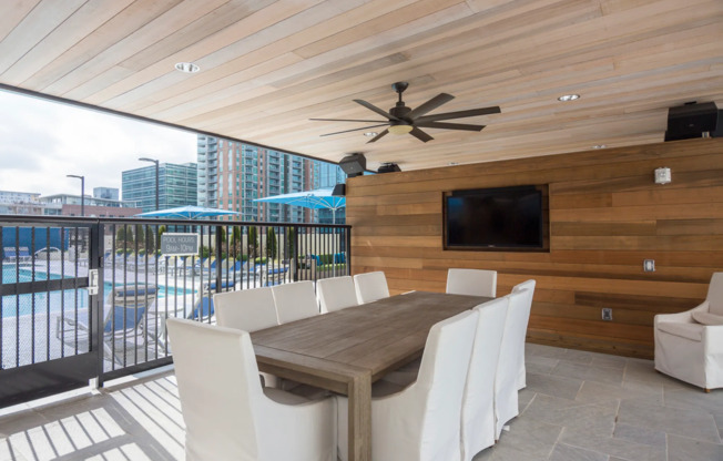 An open-air dining room by the pool enclosed with a gate, with a wood-paneled wall and ceiling, a flat-screen TV mounted on the wall, ten white upholstered chairs, and a ceiling fan.