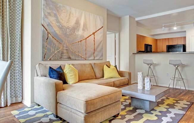 A comfortable living room here at Rio Paradiso featuring a plush sectional sofa with yellow and navy accent pillows, a modern square coffee table with decorative vases, and patterned area rug over wood-style flooring. The space opens to a kitchen breakfast bar with seating, while large wall art and soft neutral tones create a relaxed, welcoming feel.