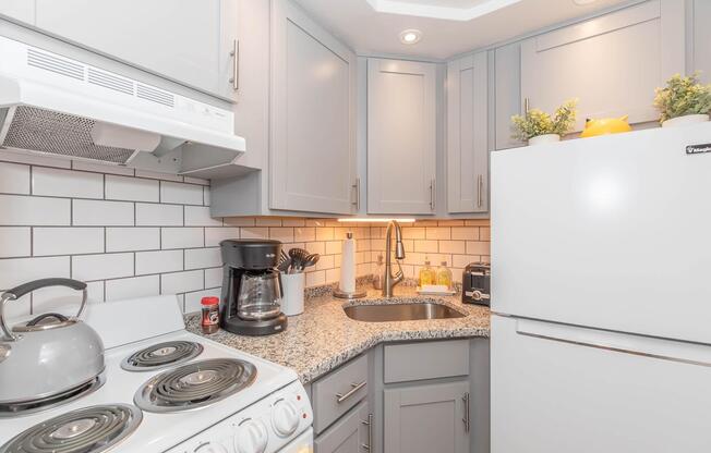 A well-organized kitchen featuring light gray cabinets, a white refrigerator, a coffee maker, and a stove. The countertops are made of speckled granite, complemented by a white tiled backsplash and small plants for decor. Bright lighting enhances the inviting atmosphere of the space.