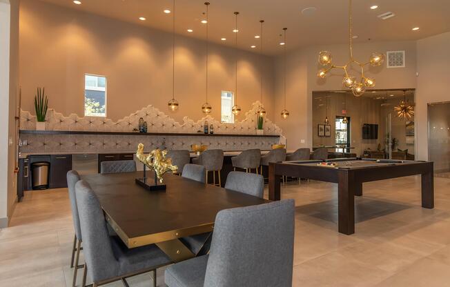 A modern communal space featuring a large dining table with gray upholstered chairs, a sleek black side table, and a game table. The room has light-colored walls, decorative wall features, and elegant hanging light fixtures, creating a stylish and inviting atmosphere.