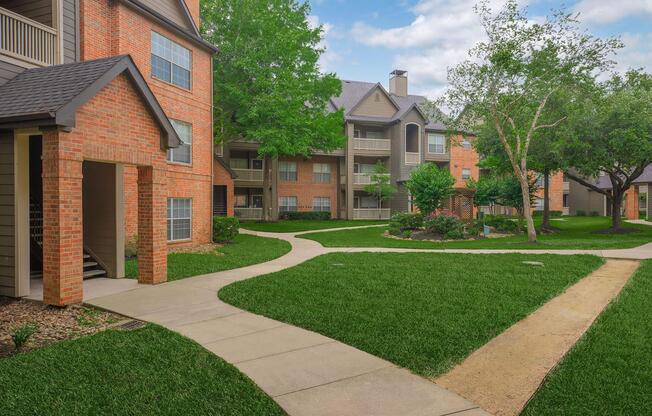 Lush green lawn with a well-maintained pathway winding through an apartment complex. Red brick buildings with balconies are visible on either side, surrounded by trees and shrubs. The sky is partly cloudy, creating a serene and inviting atmosphere.
