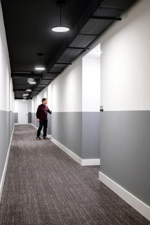 A man is standing in a hallway with grey carpeting and white walls.