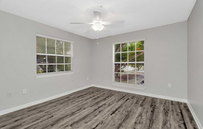 A spacious, empty room with light gray walls and a ceiling fan. Two windows with white blinds allow natural light to enter, showcasing a view of greenery outside. The floor features light brown laminate flooring, creating a clean and modern aesthetic. Ideal for personalizing or setting up living space.