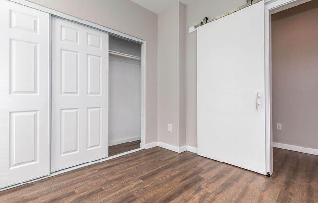 Interior of a room featuring a hardwood floor, a sliding barn door, and double closet doors. The walls are painted in a neutral color, creating a bright and spacious atmosphere.