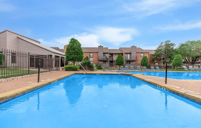 A clear blue swimming pool surrounded by a landscaped area with green trees and lounge chairs. In the background, there are multi-story apartment buildings with a neutral color palette. A fence lines one side of the pool, and the sky above is bright with a few clouds.
