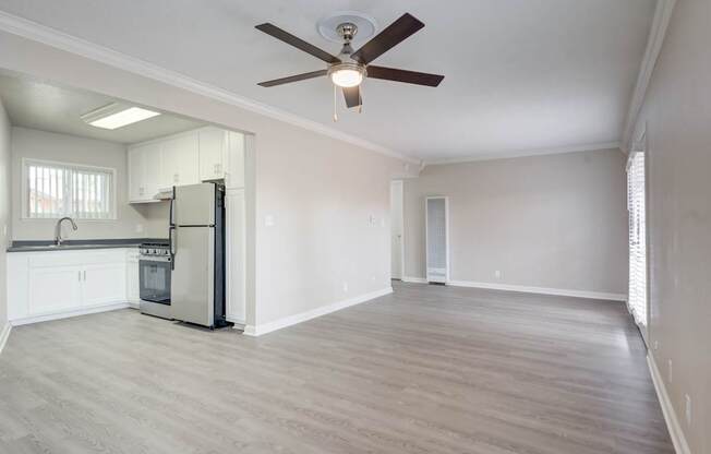 A spacious room with a ceiling fan and light fixture, a kitchen area with white cabinets and a stainless steel refrigerator, and a large window with blinds.